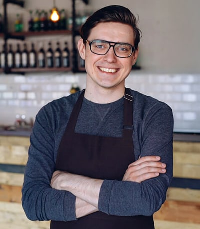 A man in an apron at a coffee shop, posing with his arms folded