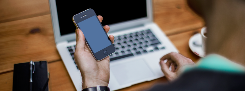 Man holding iPhone sitting at table with Macbook. 